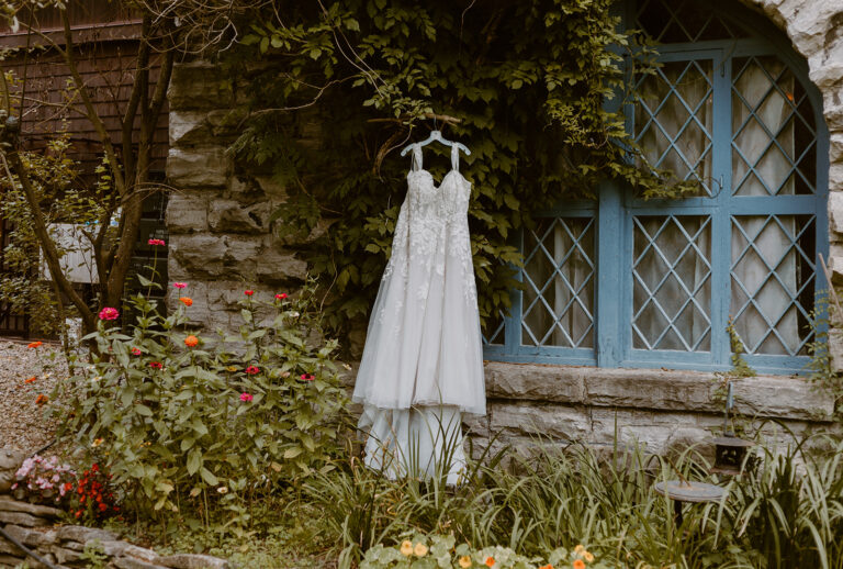 A white wedding dress hangs on a dress form in a garden beside a stone wall and a blue-framed window.
