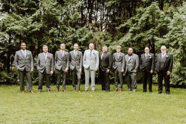 Group of men in formal suits standing in a row on a grassy lawn with trees in the background.