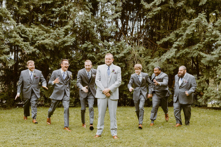 Seven men in suits posing on a grassy lawn with trees in the background.