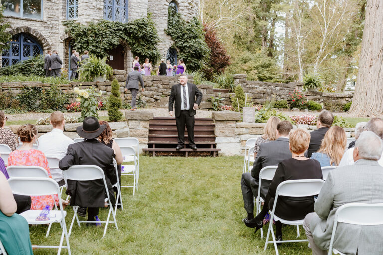 A man in a suit stands on stone steps delivering a speech at an outdoor wedding, with guests seated on the lawn.