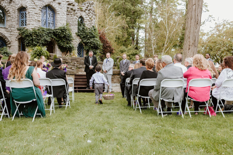 Outdoor wedding ceremony on a grassy lawn in front of a stone building covered in ivy, with guests seated on folding chairs facing the couple.