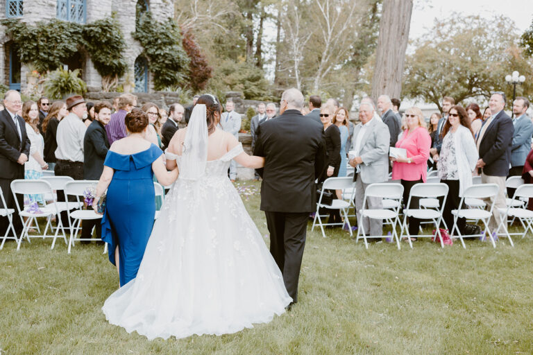 Bride and groom walk away from the camera down the aisle at an outdoor wedding, with guests seated on white chairs along a grassy lawn.