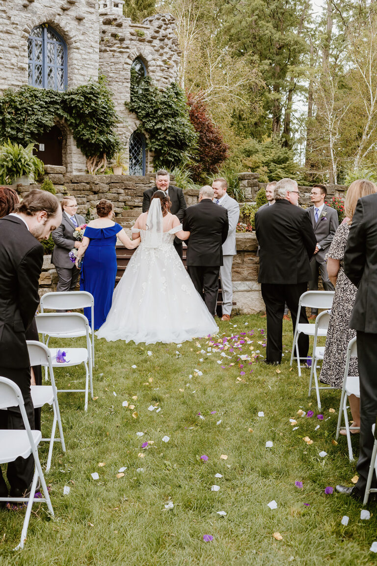 Outdoor wedding ceremony on a grassy lawn with a bride in a white gown, petals scattered on the ground, and guests seated on white chairs near a stone building.