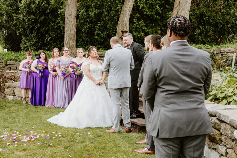 Bride and groom kiss during an outdoor wedding ceremony, with bridesmaids in purple dresses and groomsmen in gray suits standing nearby.