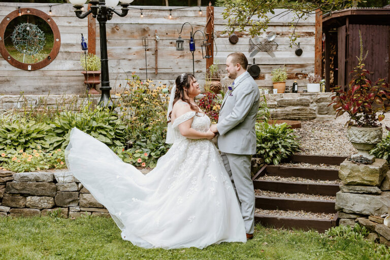 Bride in a white wedding gown with a long train and groom in a gray suit stand hand in hand on a lawn in front of a rustic wooden fence and stone steps.