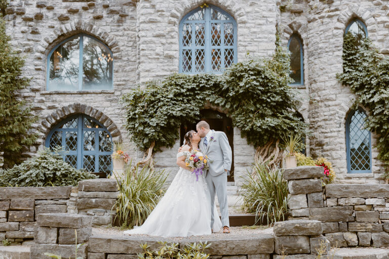 A bride in a white wedding gown and a groom in a light suit stand on a stone staircase in front of an ivy-covered stone building.