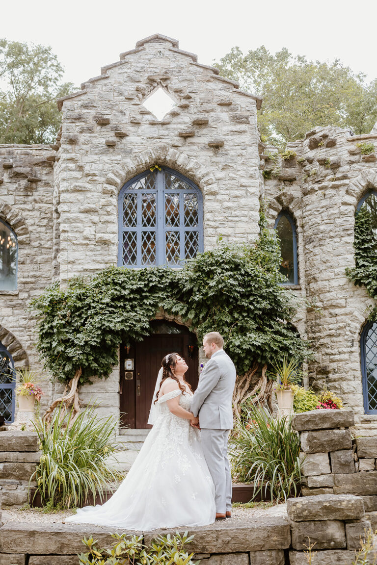 A bride and groom embrace in front of an ivy-covered stone church ruin with arched windows.
