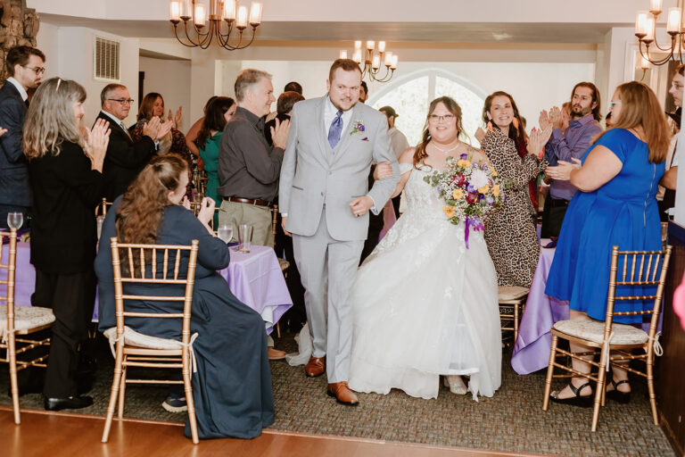 Bride in a white wedding dress and groom in a light gray suit walk through a crowd of guests applauding at a reception with purple table linens and gold chairs.