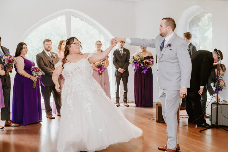 Bride and groom dance hand in hand at their wedding reception, surrounded by bridesmaids in purple dresses and other guests.