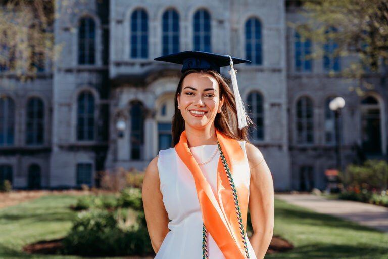 Smiling graduate wearing a cap and an orange stole over a white dress, standing on a campus lawn in front of a stone building.