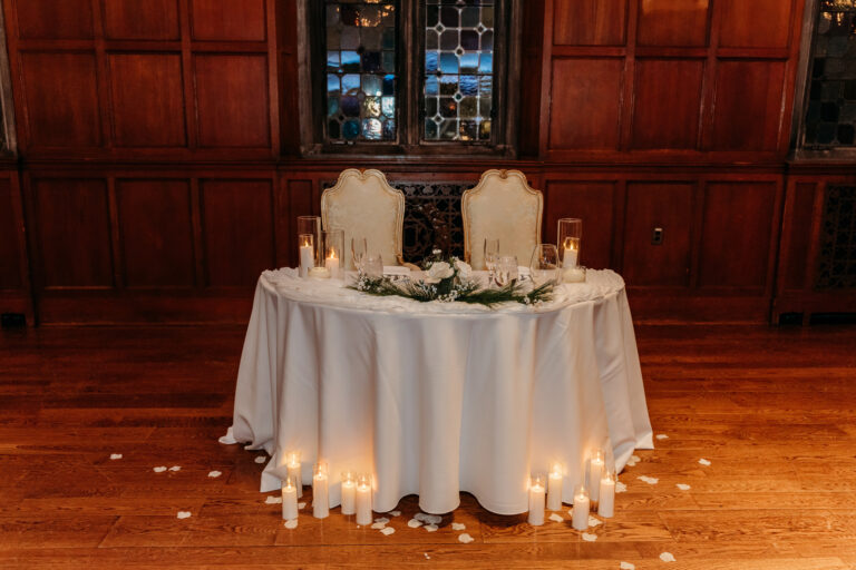 Round table with a white tablecloth, two ornate chairs, a floral centerpiece, and candles on a wooden floor in a wood-paneled room.