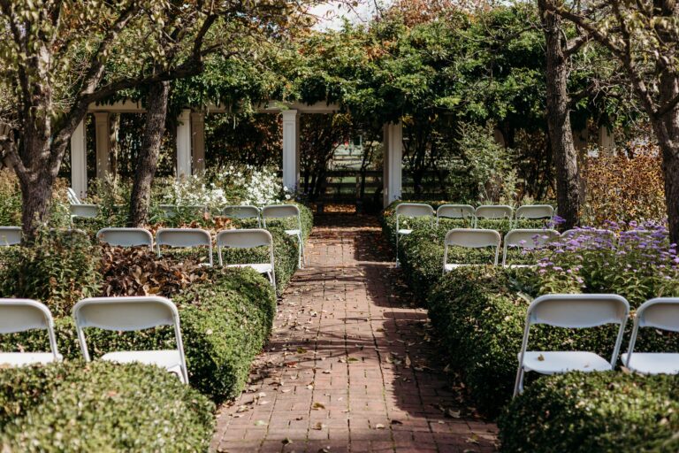 An outdoor garden aisle with a brick path, folding chairs on both sides, and neatly trimmed hedges leading to a house in the background.