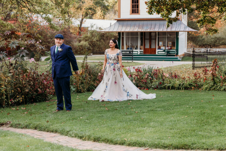 A bride in a white floral wedding gown stands on a grassy lawn beside a man in a dark suit, with a garden and house in the background.