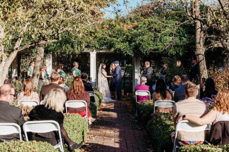 Outdoor garden wedding ceremony with the bride and groom under a wooden arbor as guests sit in rows.