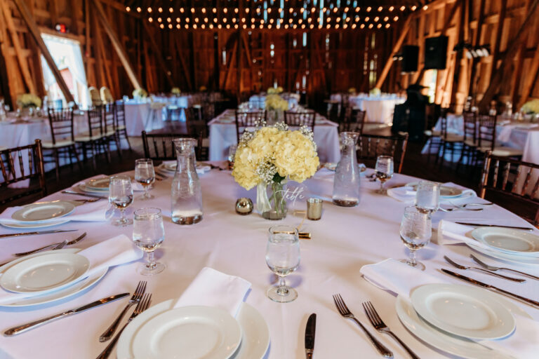 Round banquet table set for a wedding reception in a rustic wooden venue, with a white floral centerpiece and neatly arranged place settings under warm string lights.
