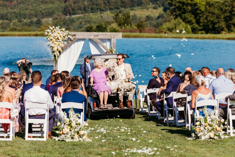 Lakeside outdoor wedding ceremony under a floral arch with guests seated on white chairs.