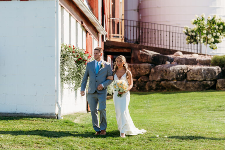 A bride in a white wedding dress and a groom in a light blue suit walk hand in hand on a sunny green lawn beside a white building.