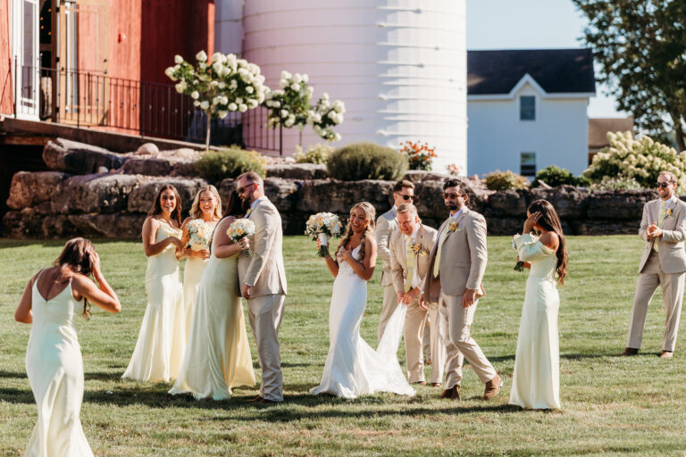 Outdoor wedding party on a sunny lawn beside a barn, with bridesmaids in pastel dresses, a bride in white, and groomsmen in light suits.