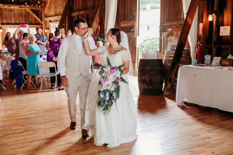 Bride in a white wedding gown and groom in a light suit walk arm in arm down a wooden barn aisle, bouquet in hand, with guests seated at tables in the background.