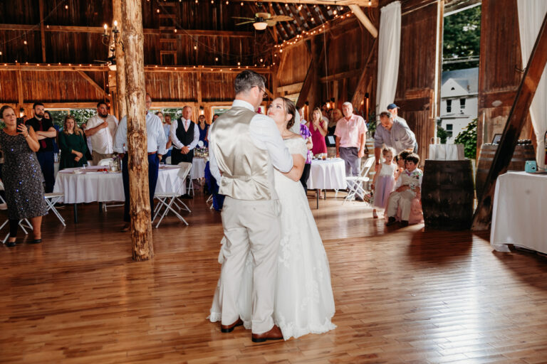 Bride in a white wedding dress and groom in a light suit dance on a wooden floor in a rustic wedding venue, with guests watching.