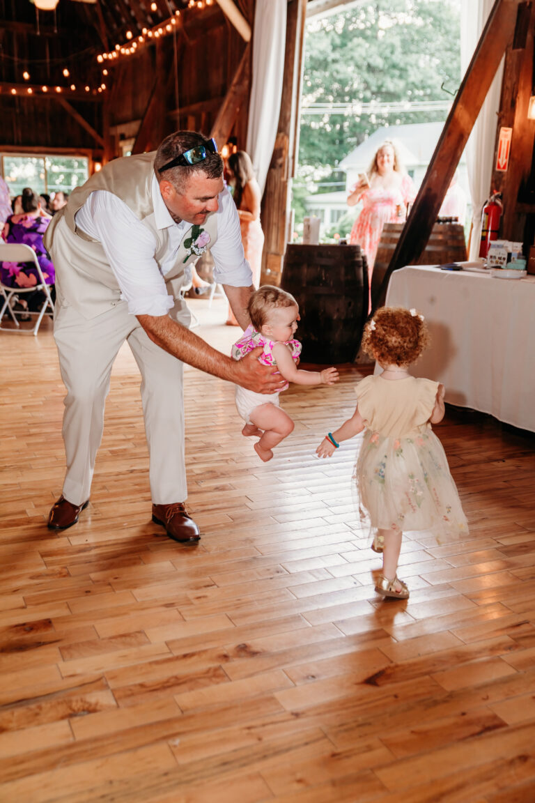 A man in light-colored clothing dances with two young children on a wooden dance floor at an indoor party.