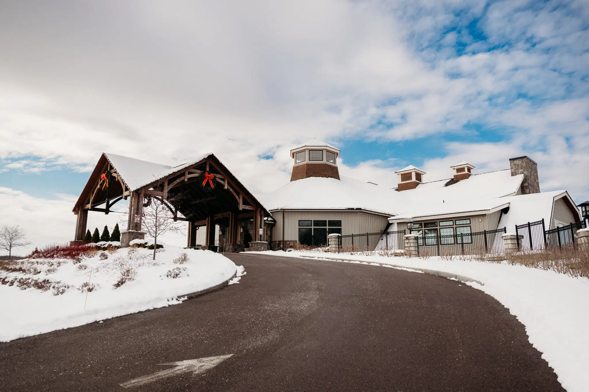 Snow-covered lodge-style building with a curved driveway and a circular tower under a partly cloudy blue sky.