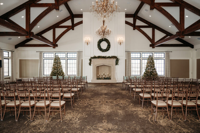A spacious event hall with exposed wooden beams, rows of chairs facing a fireplace adorned with a wreath and flanked by two decorated Christmas trees beneath a grand chandelier.