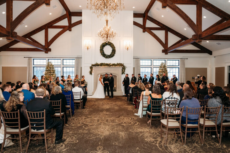 Rustic barn wedding ceremony with a couple at the altar and officiant, guests seated in wooden chairs under chandeliers, and a wreath above the fireplace flanked by Christmas trees.