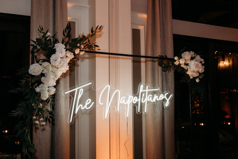 A neon sign reading 'The Napolitanos' is framed by white flowers and greenery.