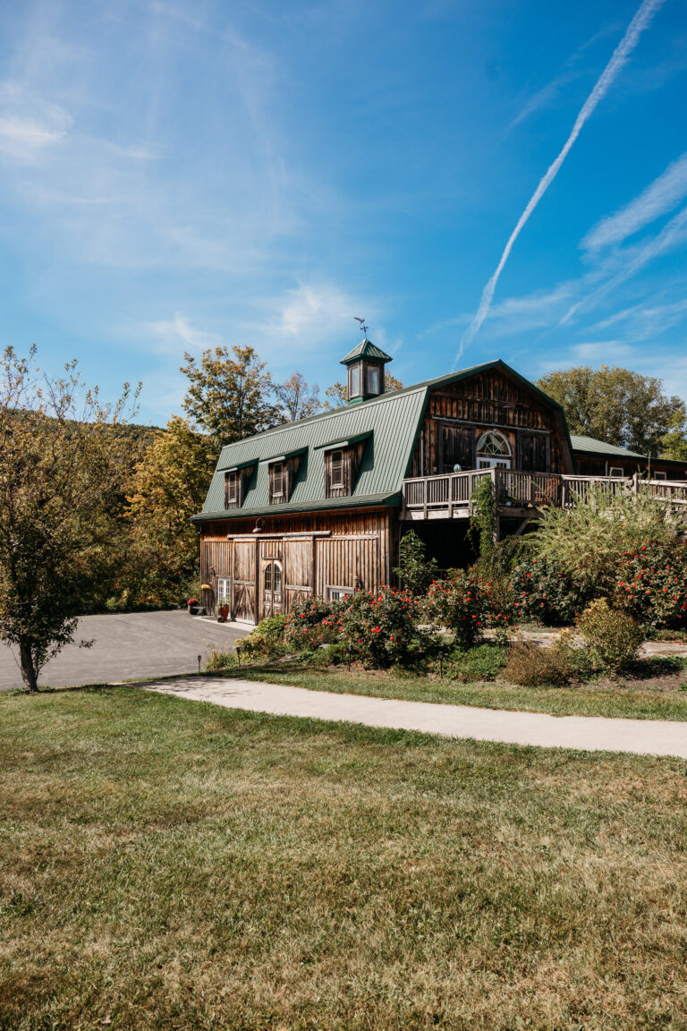 A brick-and-wood barn with a green metal roof and cupola sits on a grassy field beside a curving path under a blue sky.