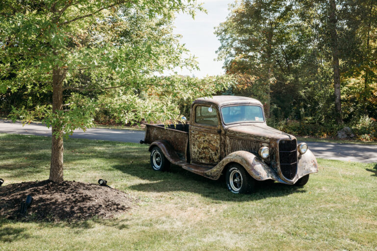 An old, rusty pickup truck sits on a grassy lawn with trees in the background.