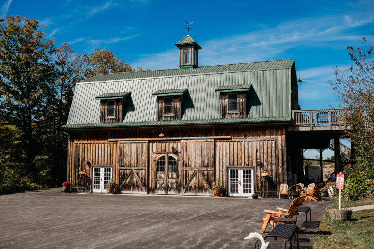 Rustic wooden barn with a green metal roof, dormer windows and a cupola, set on a gravel lot with autumn trees.