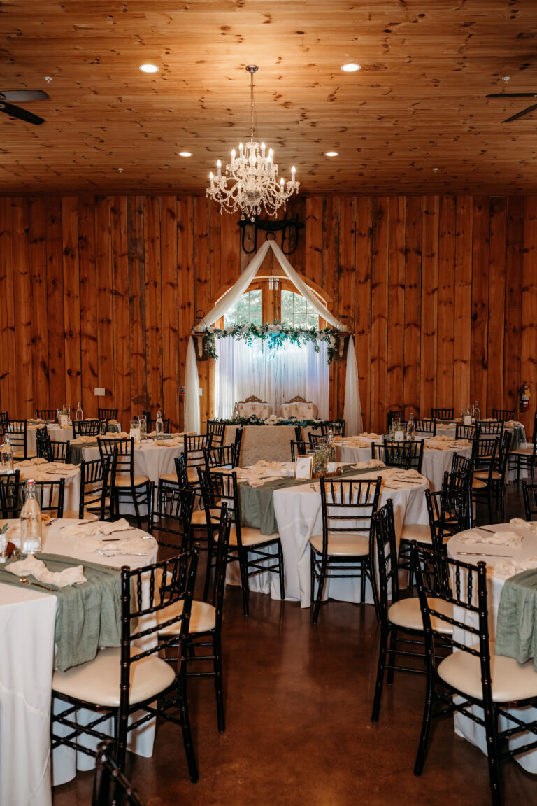 Rustic wood-paneled banquet hall arranged for a wedding reception with round tables covered in white linens, dark wooden chairs, a chandelier overhead, and a floral arch backdrop.