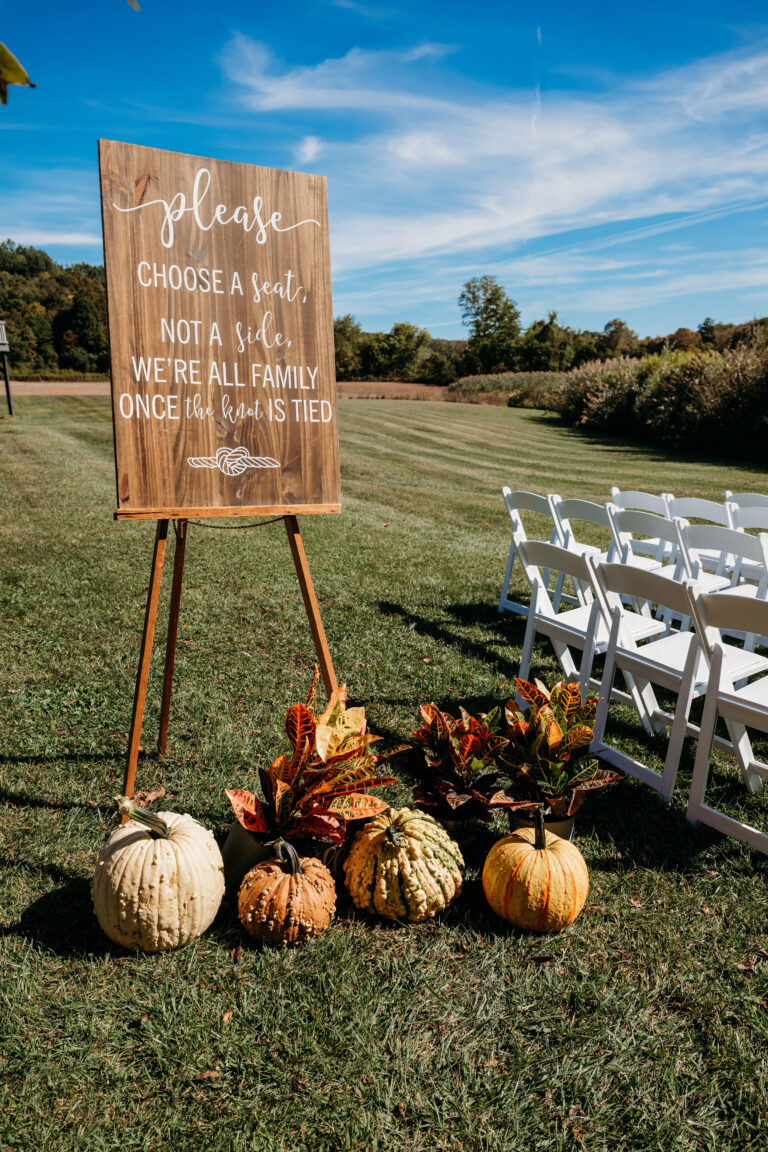 Outdoor fall event setup with a wooden chalkboard sign on an easel, pumpkins, and white folding chairs on a grassy lawn.