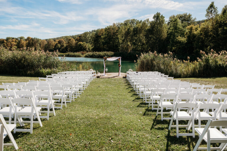 Outdoor wedding ceremony with rows of white folding chairs on a grassy lawn facing a white arch by a lake and trees.