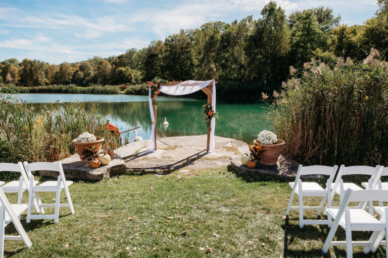 Outdoor lakeside wedding ceremony setup with a wooden arch draped in fabric and rows of white chairs on the grass.