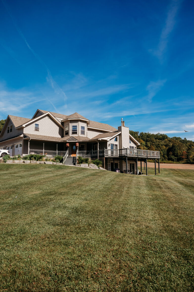A two-story suburban house with a wooden deck and a large grassy yard under a clear blue sky.