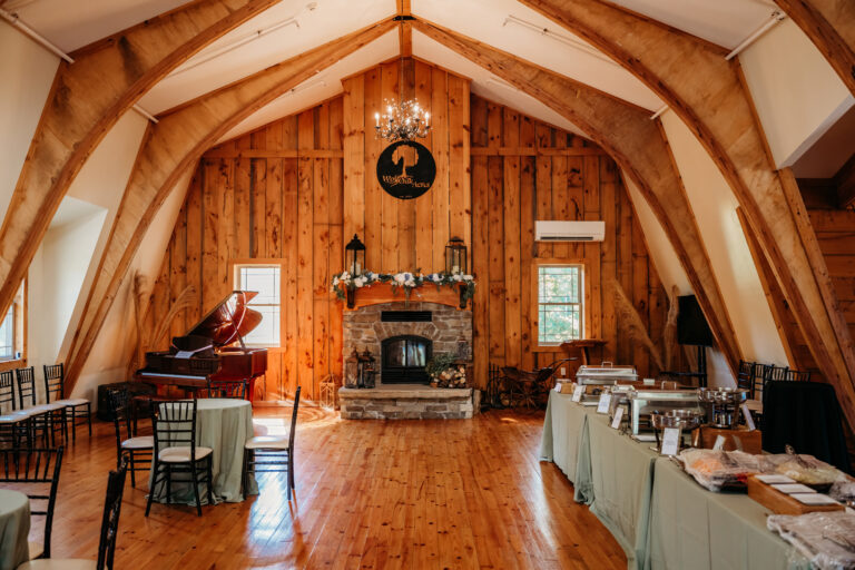 A rustic attic dining room with exposed wooden beams, a stone fireplace, a grand piano, and long tables set for an event.