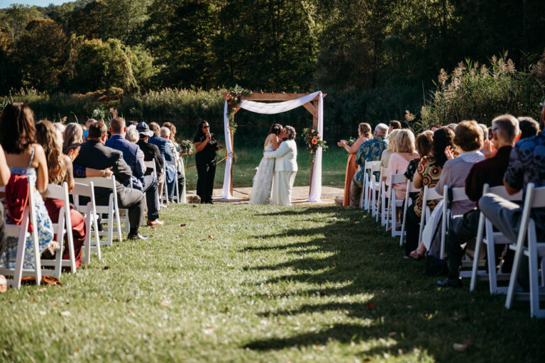 Outdoor wedding ceremony with a bride and groom under a fabric-draped arch, as guests sit on white chairs along a grassy aisle.