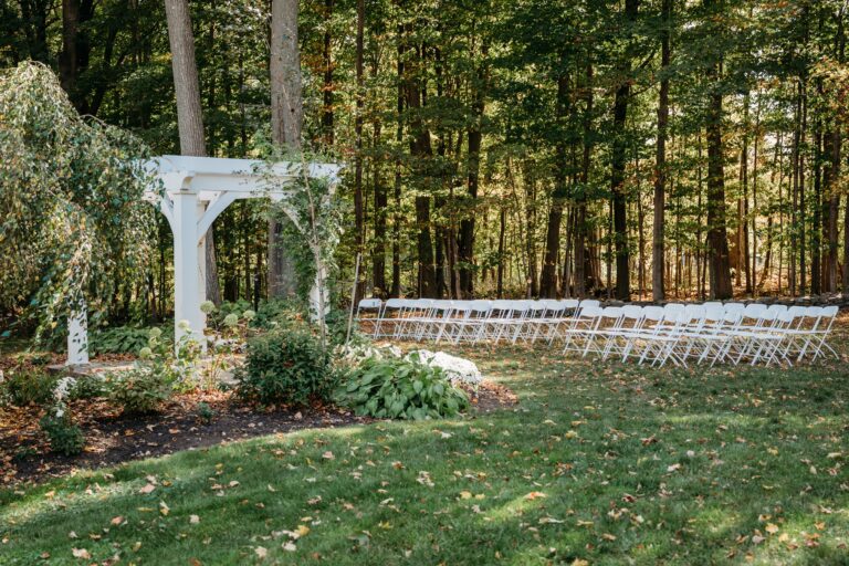 Outdoor garden ceremony space featuring a white arbor and rows of white chairs on a grassy lawn.
