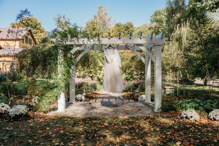 A white wedding dress hangs from a wooden garden arbor in a sunlit outdoor garden with autumn leaves scattered on the ground.