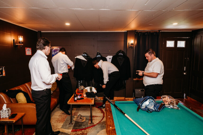 Several men in white shirts stand around a billiards table in a warmly lit basement room.