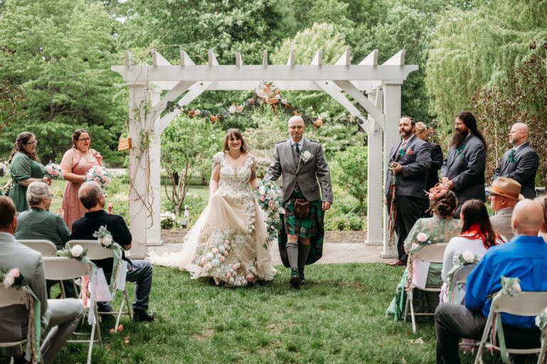 Bride and groom walk down an outdoor garden aisle under a white wooden arch, with guests seated on both sides.