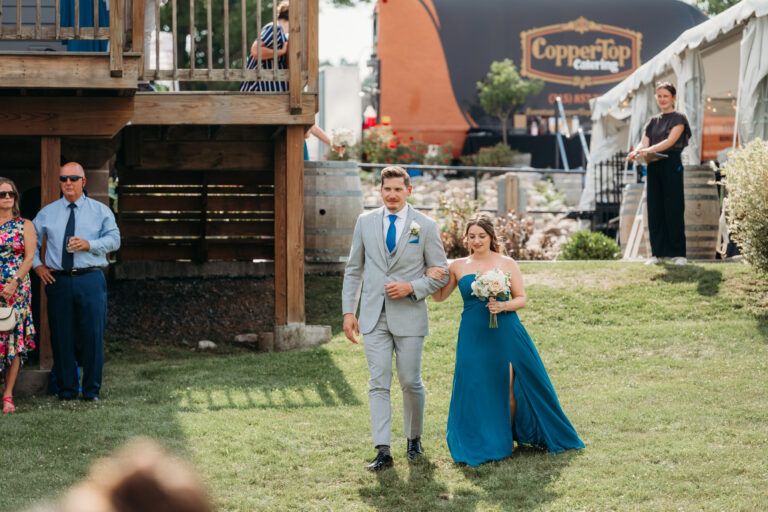 A man in a light gray suit and a woman in a blue gown walk hand in hand across a grassy outdoor wedding venue.