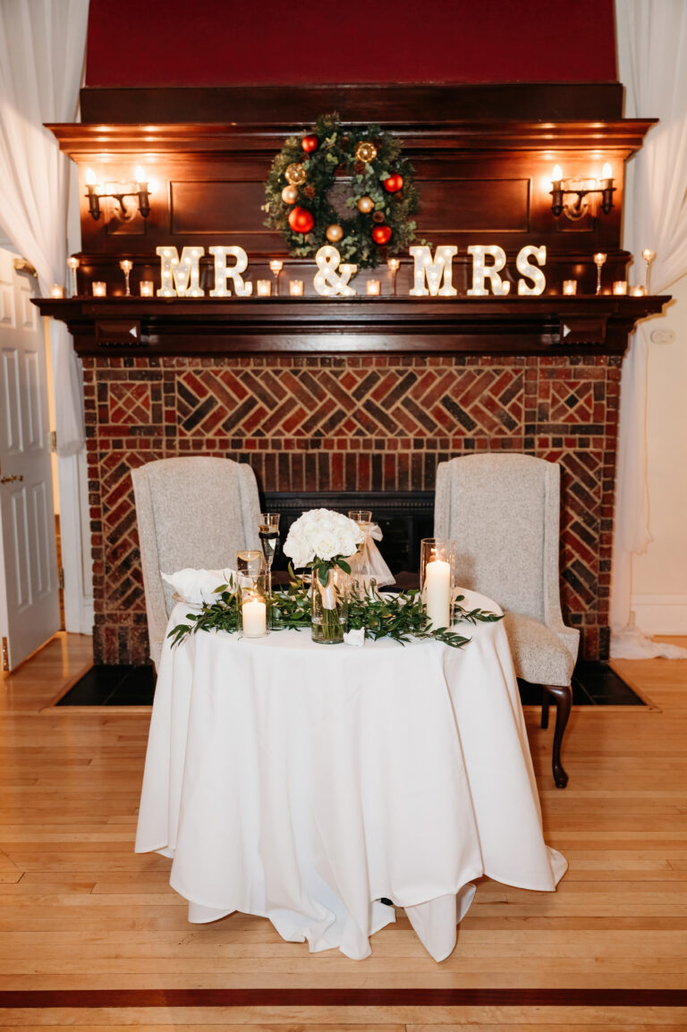 Wedding decor on a brick fireplace mantel with a wreath, candles, MR and MRS lights, two chairs, and a round table with a white cloth.