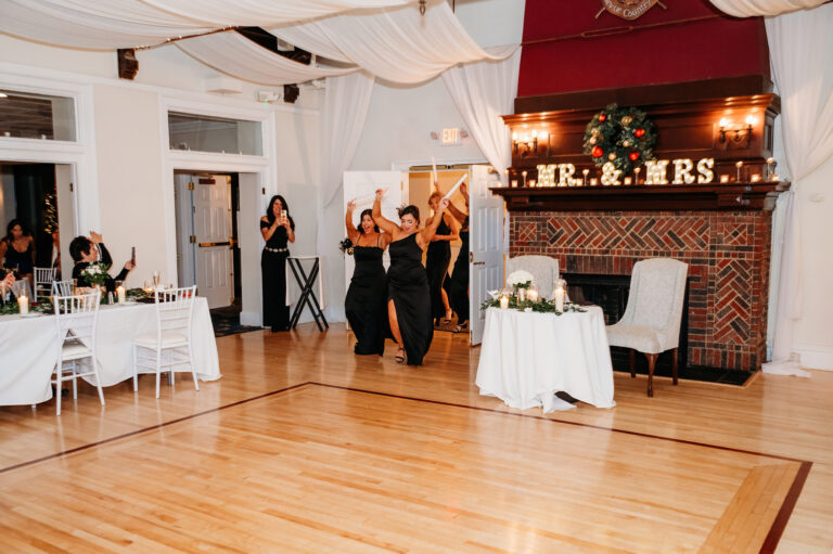 A wedding reception on a wooden dance floor with guests at tables, a couple dancing in the background, and a fireplace mantel decorated with greenery and a “MR & MRS” sign.