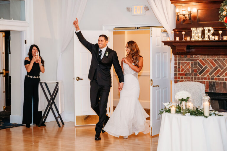 Bride in a white wedding dress and groom in a suit walk into a reception as guests applaud, with the groom raising his arm in celebration.