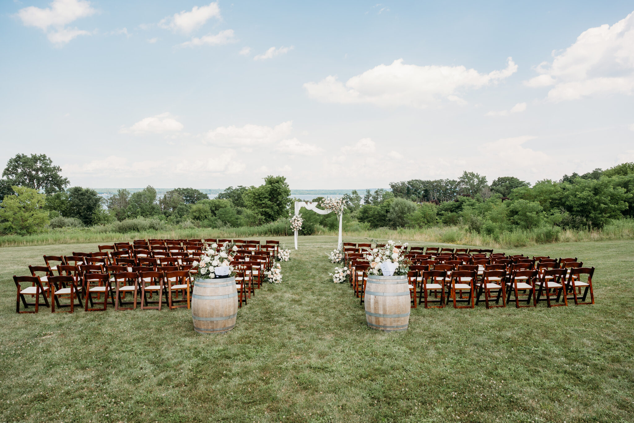 Outdoor wedding ceremony setup on a grassy field with rows of wooden chairs facing a white arch, flanked by barrels.