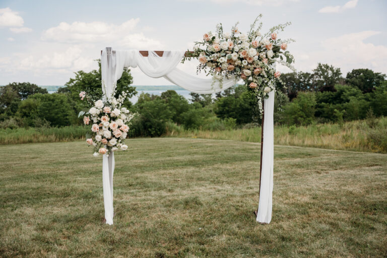 Outdoor wedding arch draped in white fabric with pink and white floral arrangements stands on a grassy field.
