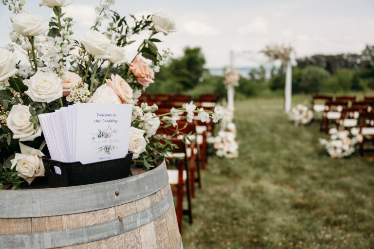 Outdoor wedding ceremony setup featuring a flower-filled wine barrel centerpiece and rows of wooden chairs on a grassy lawn.